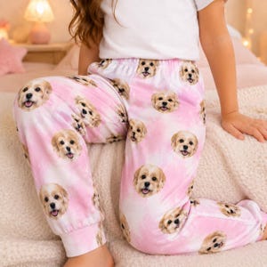 Può includere: Pantaloni da casa rosa con motivo tie-dye e una stampa ripetuta del muso di un cane dal pelo chiaro. I pantaloni presentano un elastico in vita e polsini alle caviglie, abbinati a una t-shirt bianca a maniche corte a tinta unita.