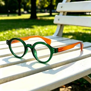 May include: Spectacles featuring round, dark green frames and translucent orange temples. The front of the frames is a solid, glossy dark green, while the arms are a contrasting, semi-transparent orange material with a smooth, polished finish.