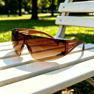 May include: A pair of wrap-around sunglasses featuring a single continuous brown tinted lens. The frame is constructed from a matching brown material with distinctive cut-out slats along the temples, providing a sleek and aerodynamic profile.