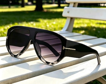 Puede incluir: Gafas de sol negras de gran tamaño con una montura gruesa y llamativa que presenta un diseño de barra superior continua. Las lentes tienen un tinte degradado oscuro y las patillas están acentuadas con un detalle metálico dorado en forma de T.