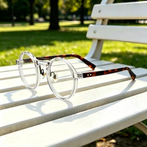May include: Round spectacles featuring a clear, transparent frame front and contrasting brown tortoiseshell patterned temples. The glasses include adjustable gold-toned metal nose pads and a thin bridge connecting the circular rims.