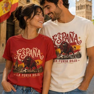 May include: A red t-shirt and a cream t-shirt, each featuring a graphic print of a bull with a soccer ball, the text ESPAÑA, and the text LA FURIA ROJA. Both shirts have short sleeves and a crew neckline, paired with blue denim jeans.