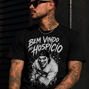 Puede incluir: Una camiseta negra de manga corta con un estampado gráfico de una persona con camisa de fuerza y una sonrisa siniestra. El texto "BEM VINDO AO HOSPICIO" está impreso en blanco sobre la ilustración, que incluye efectos de salpicaduras.