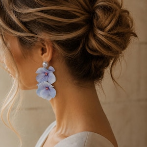 Pode incluir: Um par de brincos pendentes com um pino de pérola branca redonda no topo, ligado a duas pétalas de flor de hortênsia azul-claro sobrepostas com um pequeno detalhe rosa no centro, criando um design floral que pende do lóbulo da orelha.