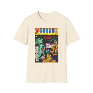May include: A cream-colored short-sleeve t-shirt featuring a graphic print of a vintage comic book cover titled Gorgo. The artwork depicts a large green reptilian monster on a rocky landscape with the text Monsters for the Moon printed in red.