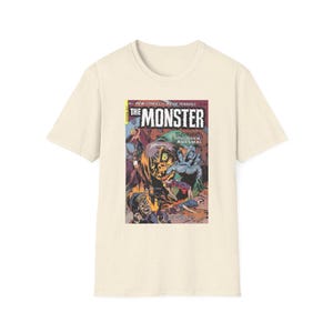 May include: A cream-colored short-sleeve t-shirt featuring a graphic print of a vintage comic book cover titled The Monster. The artwork depicts a large, monstrous creature with a green face and blue hands looming over figures in a dark scene.