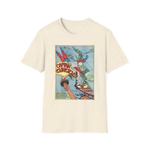May include: A cream-colored short-sleeve t-shirt featuring a vintage comic book graphic on the front. The illustration depicts a superhero flying above the Statue of Liberty's torch, with military aircraft and the text Captain Courageous.