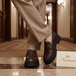 May include: Dark brown leather penny loafers with a thick black lug sole. The shoes feature a classic strap across the vamp and a smooth finish. A small white card with the gold text Noorinae and a crown icon is placed next to the footwear.