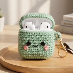 Peut inclure: Un étui de protection crocheté vert clair pour écouteurs sans fil, arborant un visage souriant avec des yeux en perles noires et de petites joues roses brodées. L'étui comprend un mousqueton en métal doré attaché sur le côté pour le transport.