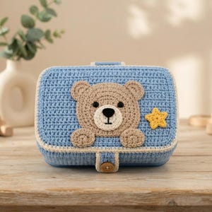 Puede incluir: Una caja rectangular de ganchillo en color azul claro con una aplicación de cara de oso en beige, dos pequeñas patas, una estrella amarilla y un cierre de botón de madera en la solapa frontal. La caja presenta un patrón de punto texturizado.