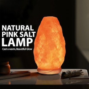 Puede incluir: Una lámpara de sal rosa natural con una textura cristalina rugosa que emite un cálido resplandor naranja. La lámpara se asienta sobre una base de madera circular. El texto a la izquierda dice: NATURAL PINK SALT LAMP. Cast a warm, Beautiful Glow.