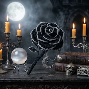 May include: A crocheted black rose with silver-toned edges on a stem, featuring a dangling silver crescent moon charm and a small dark bead. The rose is displayed alongside silver candle holders, a crystal sphere, old books, and a skull.