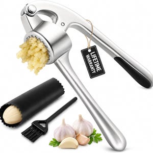May include: A silver-colored metal garlic press with black handle grips, containing crushed garlic. Includes a black silicone garlic peeler tube, a small black cleaning brush, and a black tag with the text LIFETIME WARRANTY attached by a string.