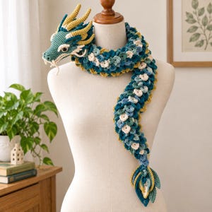 Peut inclure: Une écharpe dragon au crochet présentant une tête détaillée avec des cornes jaunes et des moustaches blanches. Le corps est composé de mailles superposées en forme d'écailles dans des tons bleu sarcelle, bleu foncé et crème, finissant en pointe.