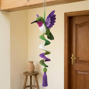 Op de afbeelding: Een gehaakte kolibrie als hangende decoratie met een groen, wit en paars lichaam en uitgestrekte vleugels. Onder de vogel bevindt zich een spiraalvormige staart van groen, wit en paars garen, afgewerkt met een paarse kwast aan de onderkant.