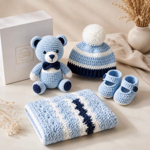 May include: A crochet set featuring a light blue and dark blue teddy bear with a bow tie, a matching striped beanie with a white pom-pom, a pair of light blue booties with buttons, and a folded striped blanket, all presented with a white gift box.