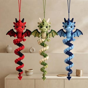 May include: Three hanging crochet dragon ornaments in red, green, and blue. Each dragon features a detailed head with horns, small wings, and a long, spiraling tail that hangs vertically. The yarn textures are visible on each creature.