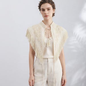 Könnte beinhalten: Ein cremeweißes ärmelloses Capelet mit detaillierter, tonaler Blumenstickerei an Schultern und Brust, verziert mit langen Fransen an den Rändern und einem dünnen Bindeband am Ausschnitt, das mit zwei hängenden Quasten versehen ist.