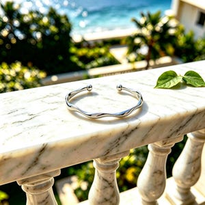 May include: A silver-toned cuff bracelet featuring a smooth, undulating wavy design. The open-ended bangle is finished with a small, polished spherical bead at each of the two terminal ends, creating a clean and reflective metallic appearance.