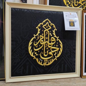 Könnte beinhalten: Ein quadratisches Wandbild mit schwarzem Stoffhintergrund und dezenter, tonaler arabischer Kalligrafie. Ein zentrales, kunstvoll goldbesticktes Emblem zeigt markante arabische Schriftzeichen. Das Werk ist in einem hellen cremefarbenen Rahmen.