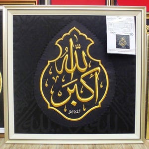 Könnte beinhalten: Ein gerahmtes Wandbild mit schwarzem Stoffhintergrund und einer großen, goldbestickten arabischen Kalligrafie in der Mitte. Die Stickerei zeigt den Schriftzug Allahu Akbar. Ein weißes Papierdokument ist in der oberen rechten Ecke befestigt.