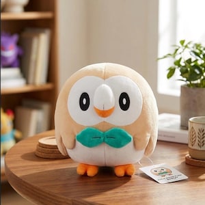 Puede incluir: Un peluche redondo de color canela y blanco con forma de búho, con un disco facial blanco, ojos ovalados negros, un pequeño pico naranja y una pajarita verde. El peluche tiene patas naranjas y una etiqueta blanca que dice Alola Region Plush Rowlet.