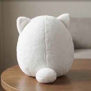 May include: A round, white plush toy with a soft, fuzzy texture. The object features two small, pointed ears at the top and a small, rounded tail at the bottom center. The back of the plush toy is smooth and uniform in its white color and rounded shape.