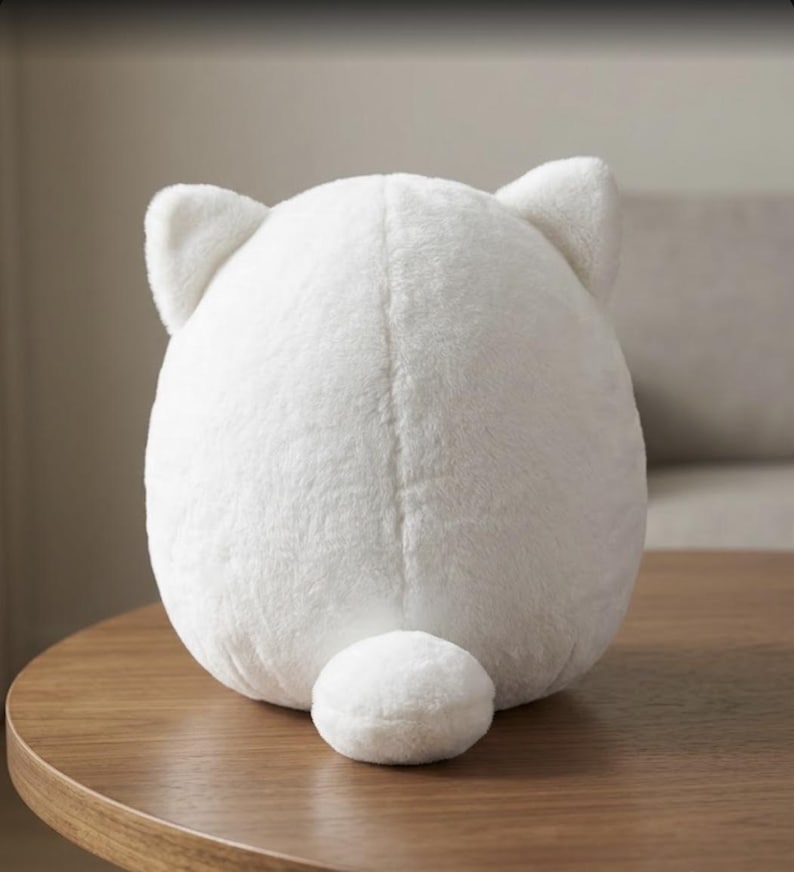 May include: A round, white plush toy with a soft, fuzzy texture. The object features two small, pointed ears at the top and a small, rounded tail at the bottom center. The back of the plush toy is smooth and uniform in its white color and rounded shape.