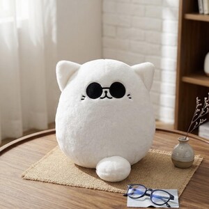 May include: A round, white plush cat toy featuring a soft, fuzzy texture, pointed ears, and embroidered black details including a small nose, mouth, and whiskers. The cat is wearing black circular sunglasses positioned above its nose.