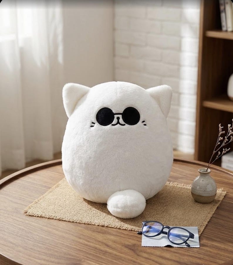May include: A round, white plush cat toy featuring a soft, fuzzy texture, pointed ears, and embroidered black details including a small nose, mouth, and whiskers. The cat is wearing black circular sunglasses positioned above its nose.