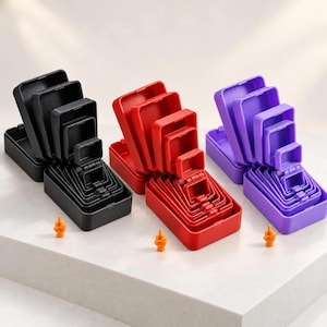 Op de afbeelding: Drie sets in elkaar passende rechthoekige doosjes in zwart, rood en paars, elk met een scharnierend deksel. Elke set bevat meerdere doosjes van afnemende grootte die in elkaar zijn gestapeld. Voor elke set staat een klein oranje figuurtje dat een middelvinger opsteekt.