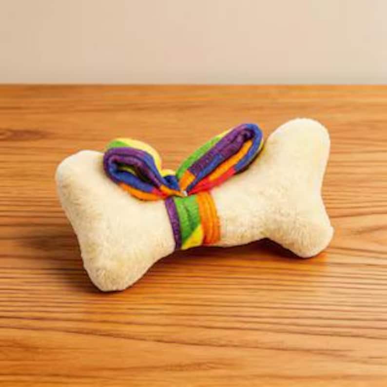 Puede incluir: Un juguete de peluche en forma de hueso de color crema con una textura suave y difusa. Una tira de tela arco&iacute;ris de colores vibrantes est&aacute; atada alrededor del centro del hueso, formando un lazo decorativo con rayas moradas, azules, verdes, amarillas, naranjas y rojas.