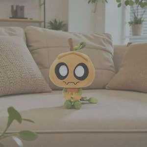 May include: A plush toy shaped like a stylized pumpkin with a light orange head, large white circular eyes with black outlines, and a small jagged mouth. The body is light green with small arms and a long, thin, curled green tail extending outward.