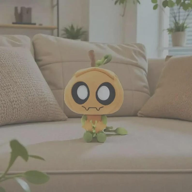 May include: A plush toy shaped like a stylized pumpkin with a light orange head, large white circular eyes with black outlines, and a small jagged mouth. The body is light green with small arms and a long, thin, curled green tail extending outward.