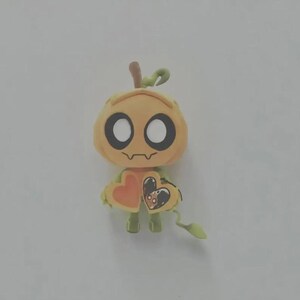 May include: A small, light orange pumpkin-shaped figurine with large white circular eyes and a thin, wavy mouth. The figure has a green stem on top and holds two heart-shaped objects in its hands, one of which features a small, detailed illustration.