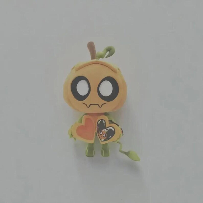 May include: A small, light orange pumpkin-shaped figurine with large white circular eyes and a thin, wavy mouth. The figure has a green stem on top and holds two heart-shaped objects in its hands, one of which features a small, detailed illustration.