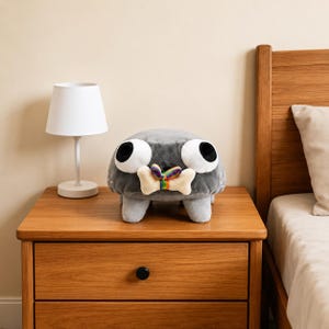 Puede incluir: Un peluche gris con forma de criatura, grandes ojos blancos y pupilas negras. El peluche tiene cuatro patas cortas y sostiene en la boca un objeto beige con forma de hueso, atado con una peque&ntilde;a cinta de colores del arco&iacute;ris.