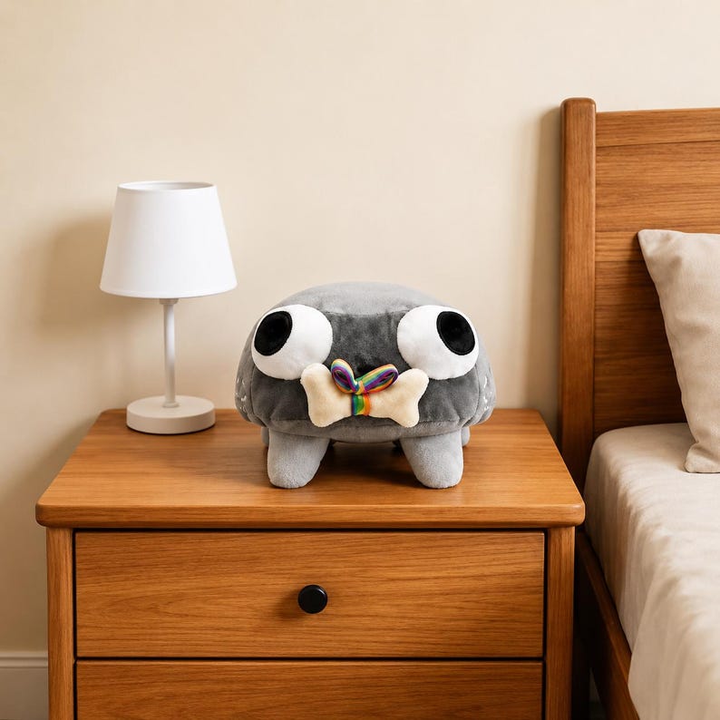 Puede incluir: Un peluche gris con forma de criatura, grandes ojos blancos y pupilas negras. El peluche tiene cuatro patas cortas y sostiene en la boca un objeto beige con forma de hueso, atado con una peque&ntilde;a cinta de colores del arco&iacute;ris.