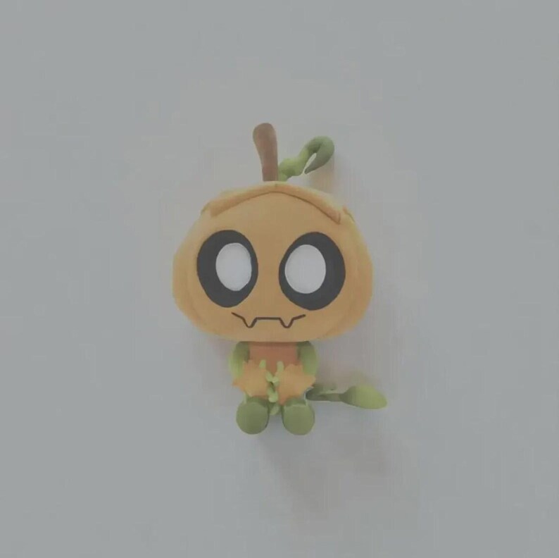 May include: A small, stylized plush figure shaped like a pumpkin with a light orange head, large white circular eyes, and a thin dark brown mouth. The body is light green with small arms and legs, and a green stem extends from the top of the head.