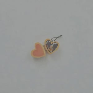 May include: A heart-shaped locket charm with a light yellow border and a black string loop. The left side is a solid peach color, while the right side features a printed illustration of a character with dark hair and large eyes inside the heart.