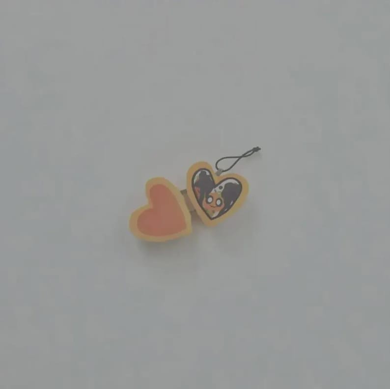 May include: A heart-shaped locket charm with a light yellow border and a black string loop. The left side is a solid peach color, while the right side features a printed illustration of a character with dark hair and large eyes inside the heart.