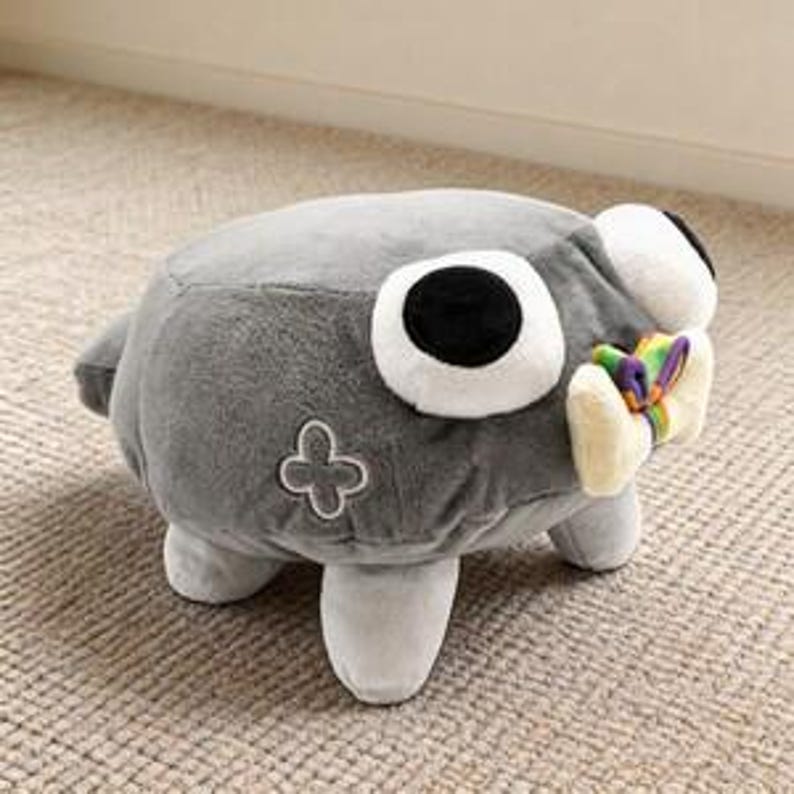 Puede incluir: Un peluche con forma de criatura gris que tiene un ojo grande de color blanco con una pupila negra. Cuenta con cuatro patas cortas, un peque&ntilde;o bordado de flor de cuatro p&eacute;talos en el lateral y un lazo color crema con rayas de colores al frente.