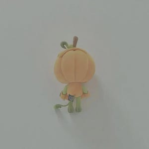 May include: A small plush toy shaped like a pumpkin with a light orange body, a green stem, and green limbs. The figure has a rounded, segmented pumpkin head, thin green arms and legs, and a small green tail. A black fabric tag is attached to the back.