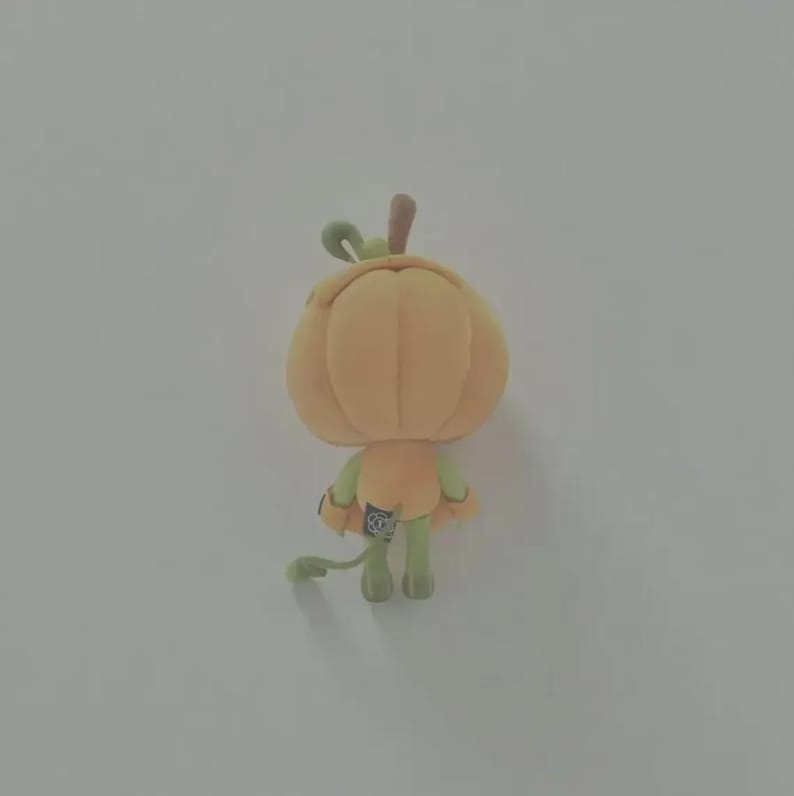 May include: A small plush toy shaped like a pumpkin with a light orange body, a green stem, and green limbs. The figure has a rounded, segmented pumpkin head, thin green arms and legs, and a small green tail. A black fabric tag is attached to the back.
