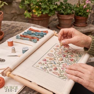 Puede incluir: Un bastidor de madera para bordar que sujeta una tela color hueso con un patrón floral de punto de cruz. El bastidor incluye un dedal de metal, una aguja y varias madejas de hilo de bordar en tonos naranja, azul y verde. El texto dice NURGE 250-2.