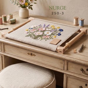 Puede incluir: Un bastidor de bordado de madera Nurge 250-3 que sujeta una tela beige con un diseño floral bordado que presenta flores rosas, amarillas, blancas y azules. El bastidor está colocado sobre un escritorio de madera con varios carretes de hilo.