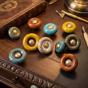 May include: A collection of round ceramic cabinet knobs featuring speckled glazes in shades of turquoise, yellow, brown, and cream. Each knob has a smooth, metallic gold-toned center button and a glossy finish, arranged on a dark wooden surface.