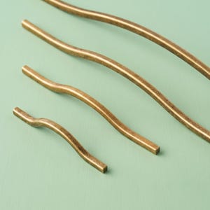 Pode incluir: Três puxadores de armário de metal com acabamento em latão antigo escovado, cada um apresentando um design ondulado e curvo distinto. Os puxadores estão dispostos em uma linha diagonal do menor para o maior, com textura metálica uniforme.
