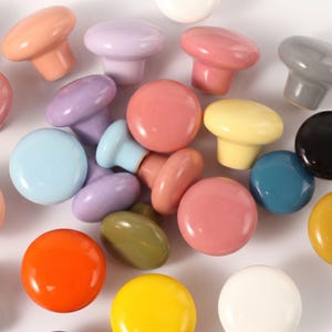 May include: A collection of round, smooth cabinet knobs in a variety of glossy colors including orange, yellow, light blue, lavender, pink, olive green, gray, black, and white. Each knob features a mushroom-shaped top and a cylindrical base.