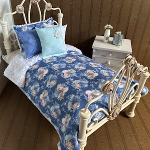 May include: A miniature white metal bed with a blue comforter featuring a princess and snowflake pattern. The bed is made up with blue and light blue pillows. A white nightstand with two drawers is next to the bed.